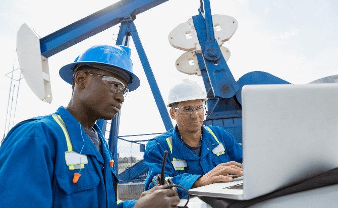 Due uomini in un campo petrolifero utilizzano un laptop mentre discutono soluzioni di Performance Management per ottimizzare l'efficienza operativa e migliorare le prestazioni nel settore energetico.